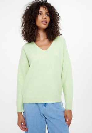 V-Ausschnitt, hellgrüner Strickpullover mit langen Ärmeln und geripptem Saum. Kombiniert mit hellblauen Denimhosen. Weiche Textur, lockere Passform.
