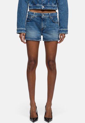 Femme portant une veste en denim bleue assortie et un short avec des détails scintillants subtils, associés à des talons hauts noirs à bouts pointus.