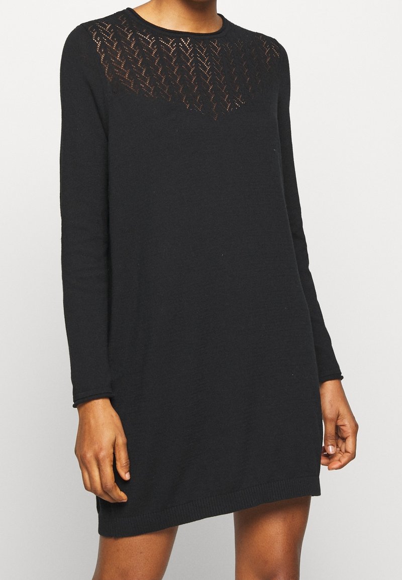Femme portant une robe tricotée noire à manches longues avec un panneau transparent à motifs sur la partie supérieure de la poitrine et les épaules.