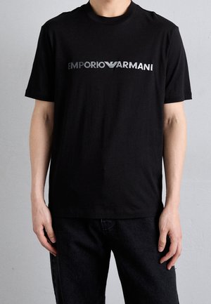 Mężczyzna w czarnej koszulce z krótkim rękawem z srebrnym logo "Emporio Armani" na środku klatki piersiowej na jednolitym szarym tle.