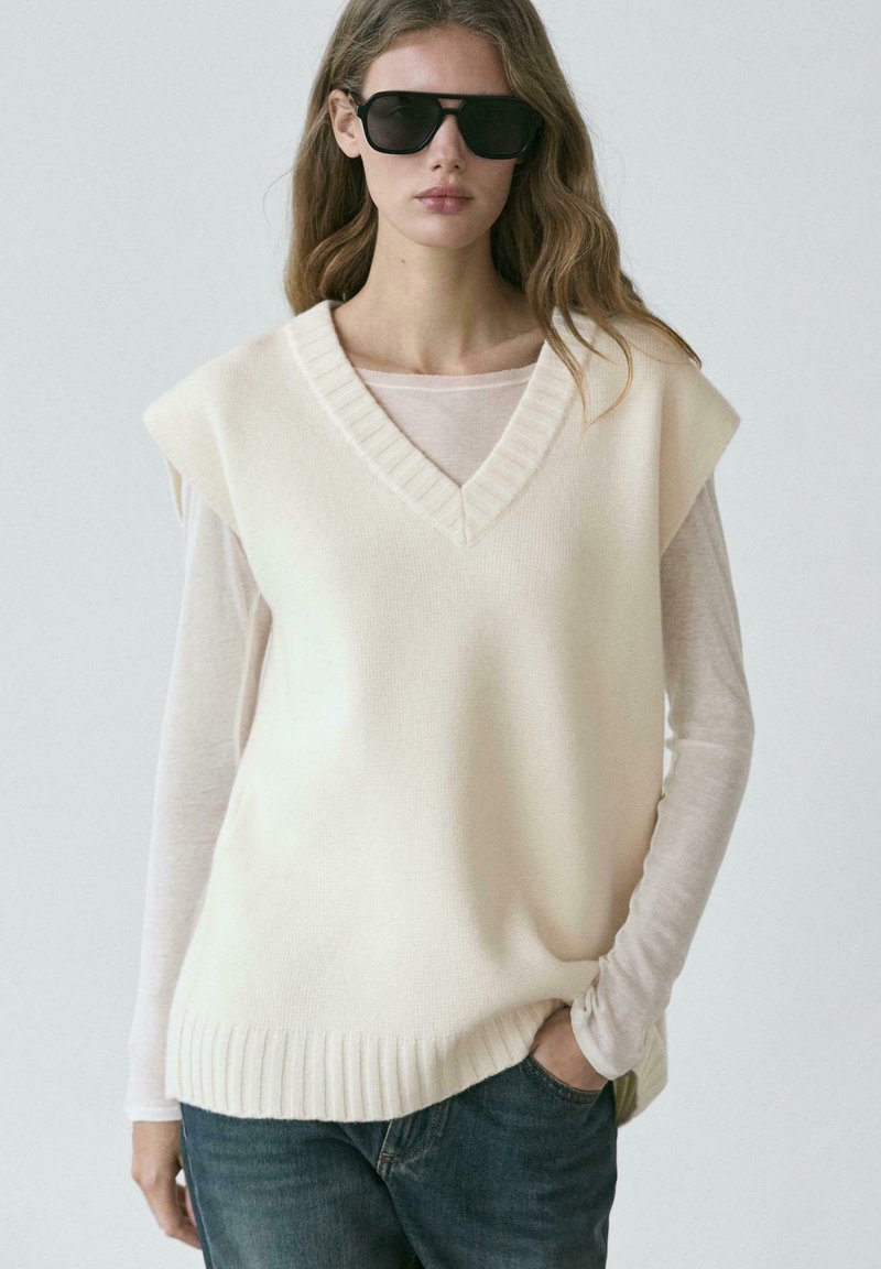 Femme portant un pull sans manches en tricot crème oversize sur une chemise blanche transparente à manches longues, lunettes de soleil foncées, et jean bleu, main dans la poche.