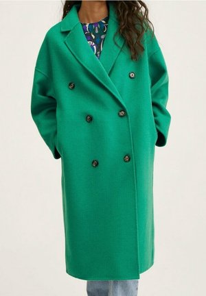 Femme portant un manteau en laine vert vif à double boutonnage par-dessus un haut à motifs et un jean bleu clair, les mains dans les poches du manteau.