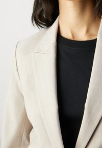 Kobieta ubrana w beżową marynarkę na czarnej bluzce z okrągłym dekoltem, widoczna od szyi do połowy torsu na jednolitym tle.