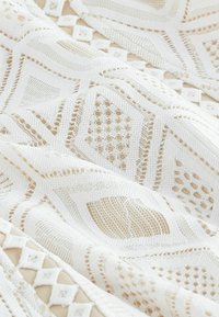 Tela de punto blanca con patrones texturizados, que presenta formas geométricas y tejidos abiertos. El material es suave con un diseño detallado.