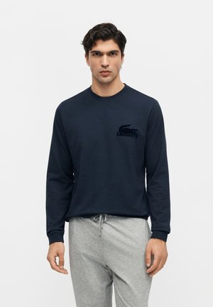 Nuori mies, jolla on tummat hiukset, pukeutunut tummansiniseen pitkähihaiseen Lacoste-paitaan ja vaaleanharmaisiin collegehousuihin, seisoo yksiväristä valkoista taustaa vasten.