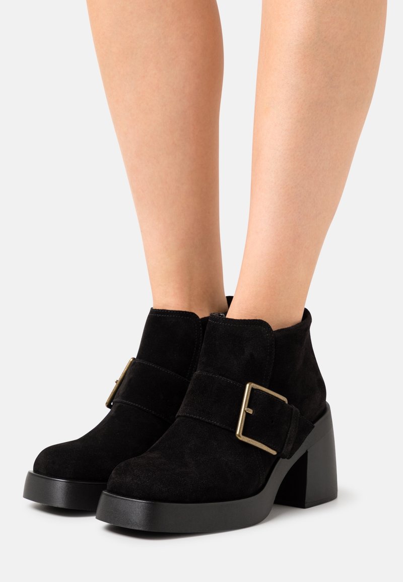 Bottines en daim noir avec des talons blocs épais et des larges sangles munies de grandes boucles dorées, portées sur des jambes nues.