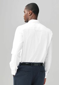 Chemise blanche à manches longues avec un col classique, coupe ajustée et poignets à boutons, accompagnée de pantalons texturés foncés et d'une ceinture noire.