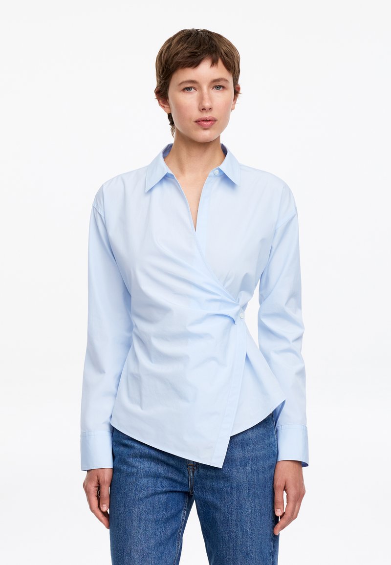 Femme aux cheveux courts marron portant une chemise bleu clair à boutons de style portefeuille et un jean bleu, faisant face vers l'avant sur un fond blanc.