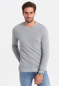 Hellegrau gestrickter Pullover mit rundem Ausschnitt, gerippten Bündchen und Saum, getragen über blauen Jeans. Der Stoff hat eine strukturierte Oberfläche.