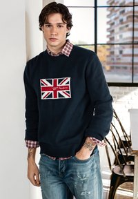 Navy sweater with a knitted Union Jack patch, paired with a red checkered shirt and distressed blue jeans. Displaying tattoos on the wrist.