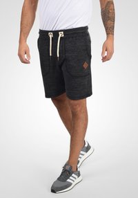 Pantalones cortos deportivos de color gris oscuro con una tela texturizada, cintura elástica, cordón blanco, bolsillos laterales y un pequeño parche de cuero en forma de diamante.