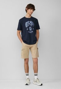 Marineblauwe katoenen T-shirt met grafische print, gecombineerd met beige cargoshorts. Accessoires zijn zwart-wit gestreepte sokken en lichte sneakers.