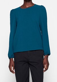 Maglietta a maniche lunghe color teal con scollatura rotonda, tessuto testurizzato e maniche arricciate, abbinata a pantaloni cargo neri.