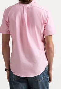 Kortärmad rosa skjorta med knappar och krage, bakveckdetalj och rundad nederkant, tillsammans med blekta blå jeans.