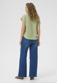 Top corto a maniche corte verde con texture e orlo curvo, abbinato a jeans blu a gamba larga e scarpe marrone chiaro.
