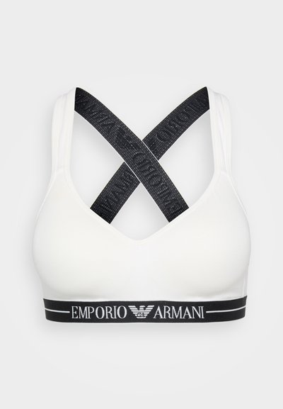 White sports bra with a black elastic band featuring "Emporio Armani" logo. Crossed black straps create a distinct design. Smooth texture.