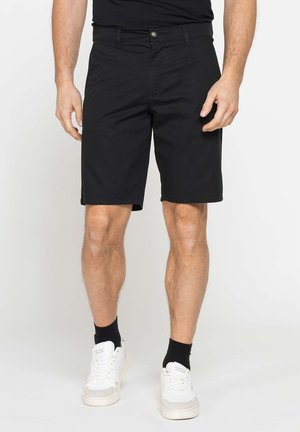Uomo che indossa pantaloncini neri al ginocchio, calzini neri e scarpe da ginnastica bianche, in piedi davanti a uno sfondo semplice.