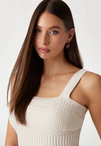 Gehaakte tanktop in crème met een vierkante halslijn, gestructureerde kabeldetails en brede bandjes. Gouden en rode oorknoppen accentueren de look.