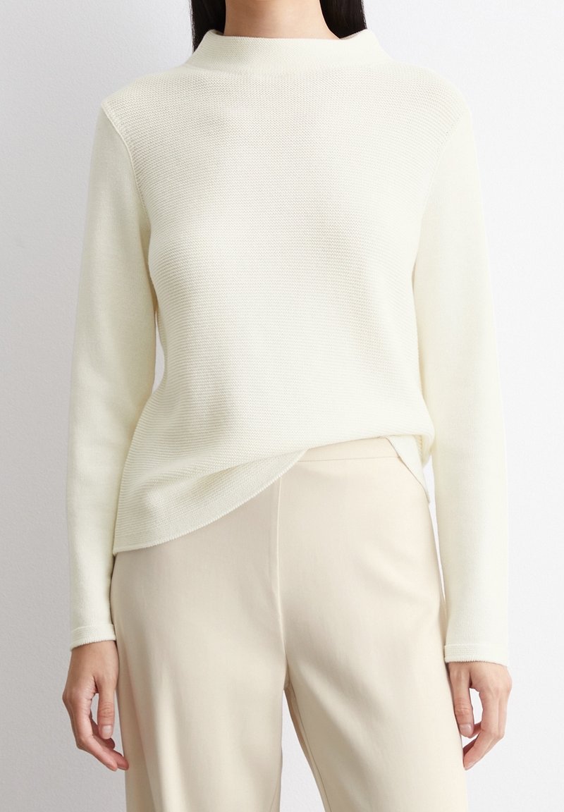 Maglione a maniche lunghe di colore crema con scollo alto e vestibilità rilassata, abbinato a pantaloni beige. Il maglione presenta un motivo testurizzato.