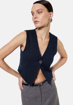 Vrouw in een mouwloos marineblauw pinstripe gilet met één knoop, gecombineerd met een grijze broek en een zwarte riem, kijkt opzij.