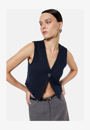 Vrouw in een mouwloos marineblauw pinstripe gilet met één knoop, gecombineerd met een grijze broek en een zwarte riem, kijkt opzij.