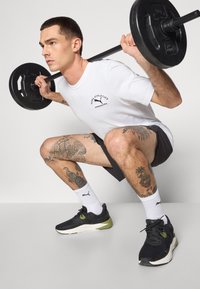 Sorte atletiske sneakers med grønne accenter, hvide logo-sokker, en hvid T-shirt med sort logo og grå shorts. Person udfører en squat.