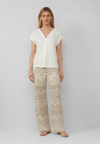 Witte blouse met een V-hals en korte mouwen, gecombineerd met beige wijde broek met zigzagpatroon. Sandalen maken de outfit compleet.