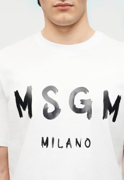 Torse masculin portant un T-shirt blanc avec l'inscription « MSGM MILANO » en style coup de pinceau noir imprimée sur la poitrine.