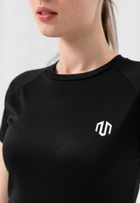 Svart kortermet treningstopp laget av glatt stoff, med en hvit logo på brystet og en tettsittende design.