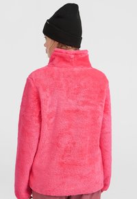 Roze fleecejack met hoge kraag, zachte textuur en lange mouwen; gedragen met een zwarte beanie. Achteraanzicht toont de helderheid van de stof.