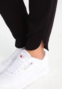 Černé zúžené kalhoty s bočními rozparky u kotníků, doplněné bílými nízkými teniskami s červeným a šedým logem.