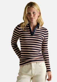 Jauna sieviete ar blondiem matiem, ģērbta jūras zila, baltā un sarkanā svītrainā garā piedurkņu polo krekla un gaismas krāsas biksēs, stāv ar vienu roku aiz muguras.