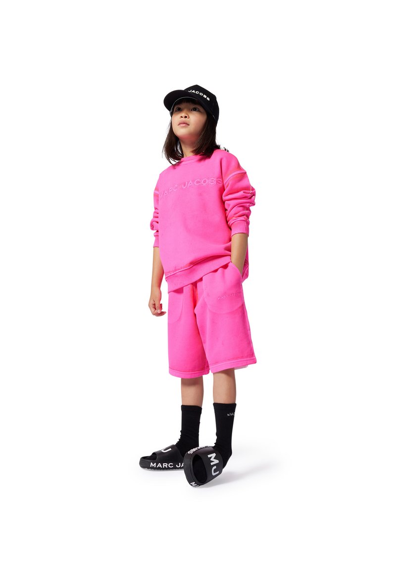 Felpa e pantaloni corti rosa neon con una texture morbida, caratterizzati da una stampa a rilievo. Un cappellino nero e sandali a infradito completano l'outfit.