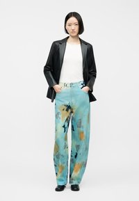 Γυναίκα με ίσια μαύρα μαλλιά, φορώντας λευκό πουκάμισο, μαύρο δερμάτινο σακάκι, φαρδιά παντελόνια tie-dye σε ανοιχτό μπλε χρώμα και μαύρα παπούτσια, στέκεται με τα χέρια στις τσέπες.