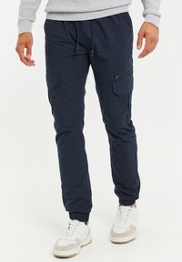 Marine cargobroek met elastische tailleband, koord, zijzakken en ritszakdetails. De stof heeft een gladde, lichte textuur.