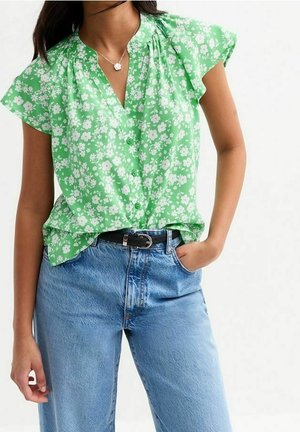 Žena nosící zelenou, krátký rukáv, květinovou košili na zapínání s světle modrými džínami s vysokým pasem a černým pásem, ruka v kapse.