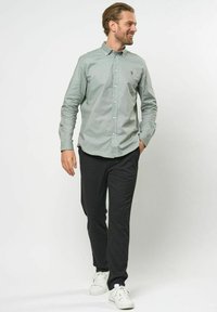 Hellgrüne Button-up-Hemd mit Kragen und gesticktem Logo, kombiniert mit dunkelgrauen, schmal geschnittenen Hosen und weißen Sneakers. Glatte Stofftextur.