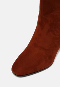 Gros plan sur une botte cheville en daim couleur rouille avec un bout pointu et des coutures visibles sur le côté.