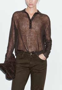 Femme portant un polo à manches longues transparent marron, rentré dans un pantalon taille haute marron foncé, tenant une pochette en daim marron assortie.