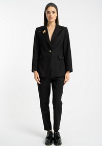 Czarna dopasowana marynarka z głębokim dekoltem w serek, z jednym złotym guzikiem i dwiema przednimi kieszeniami. W połączeniu z dopasowanymi czarnymi spodniami i czarnymi butami.
