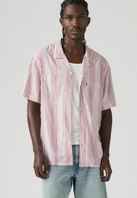 Camicetta a maniche corte bianca con strisce verticali rosse, dotata di colletto, due tasche frontali e una vestibilità rilassata, abbinata a jeans in denim chiaro.