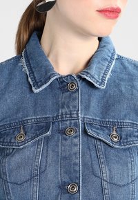 Jeansjacka i ljusblått, med krage, fransade kanter, två bröstfickor och silverfärgade knappstängningar.