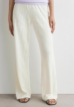 Pantalon blanc à jambes larges avec de subtiles rayures verticales porté par une personne debout, associé à des sandales noires à bout ouvert et un haut lilas.