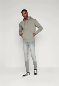 Άνδρας με τα χέρια στις τσέπες, φορώντας γκρίζα κουκούλα, ανοιχτό γκρι τζιν και μαύρα-άσπρα sneakers, στέκεται μπροστά σε απλό ανοιχτό φόντο.