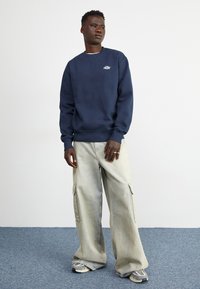 Felpa blu navy con colletto rotondo e polsini a coste, abbinata a pantaloni cargo grigio chiaro e sneaker con dettagli grigi. Finiture in tessuto testurizzato.