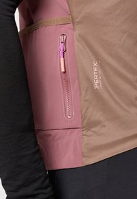 Pertex jakna s temnim črnim rokavom, rjavim telesom in roza poudarki. Vključuje zadržno žep z vijoličnim potegom in tekstovno blagovno znamko.