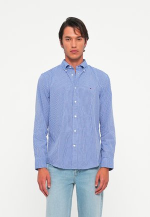 Man in een blauwe geruite overhemden met lange mouwen en lichtblauwe jeans, staand tegen een effen witte achtergrond.
