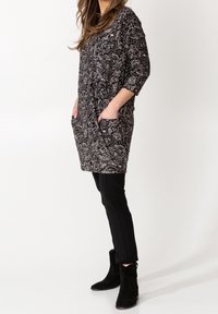 Kvinna klädd i en tunika med svart och vit paisleymönster och fickor, svarta smala byxor och svarta mocka ankle boots, stående på sidan.