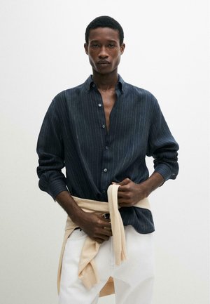 Mann trägt ein schwarzes Nadelstreifenhemd über weißer Hose und bindet einen beigen Pullover um seine Taille vor schlichtem hellem Hintergrund.