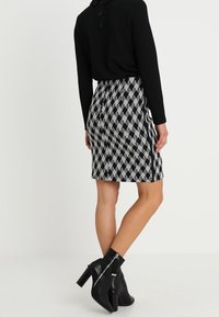 Suéter negro con botones combinado con una falda de longitud hasta la rodilla que presenta un patrón de diamantes en blanco y negro, acompañado de botas negras hasta los tobillos.